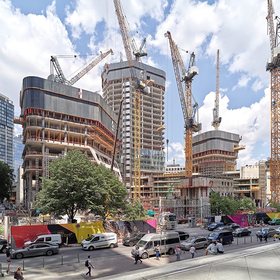 Die Konstruktion der Hochhäuser mit Gerüsten und Kränen. Davor eine befahrene Straße und belebte Fußgängerzone.