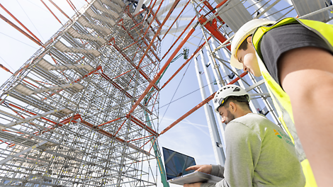 Zwei Männer auf der Baustelle. Einer hebt einen Laptop, sie schauen auf ein Gerüst.