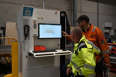 Two men in high-vis are looking at a screen on the side of a cutting machine,