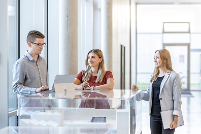 Three people in business attire stand around a desk and a laptop.