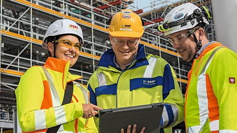 Zuverlässige Dienstleistungen in allen Projektphasen Drei Menschen in neonfarbener Arbeitsschutzkleidung stehen vor einer eingerüsteten Baustelle und schauen gemeinsam auf ein Tablet.