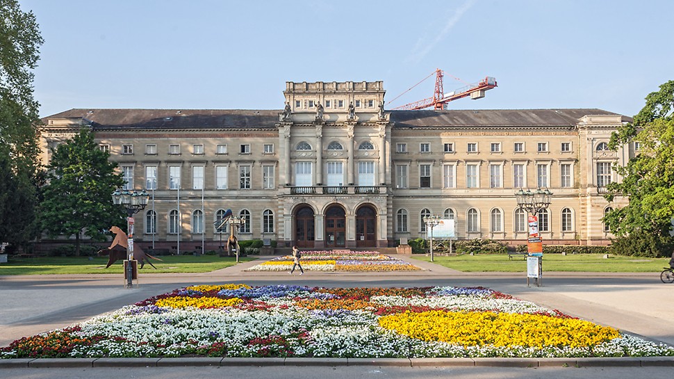 Front des Karlsruher Naturkundemuseums. Während auf dem Weg im Vordergrund eine Passantin läuft, kann man im Hintergrund einen Kran hinter dem Gebäude aufragen sehen. 